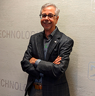 Smiling man with gray hair in a blazer, arms crossed, standing in front of a wall with the word TECHNOLOGY visible.